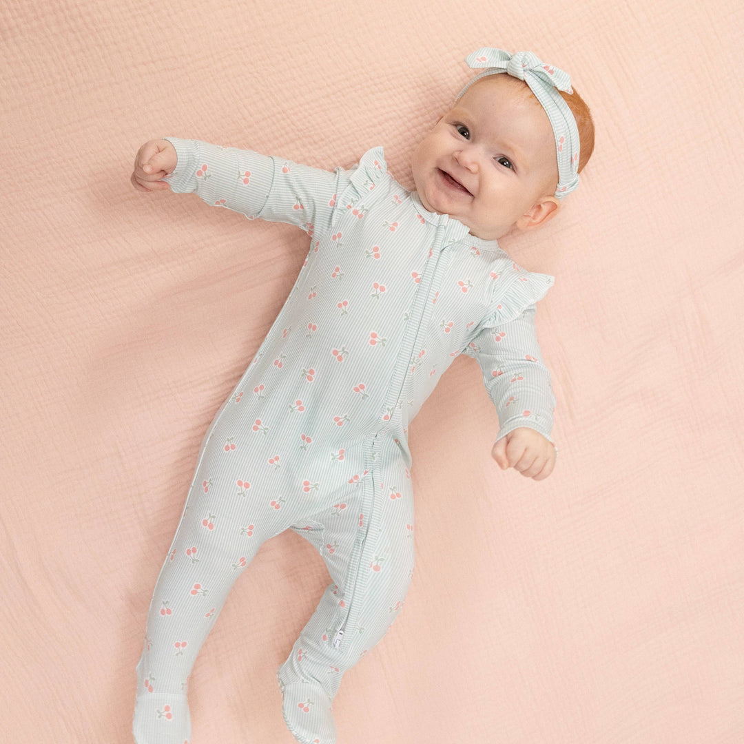A smiling baby lies on a pale pink blanket, wearing Angel Dear&