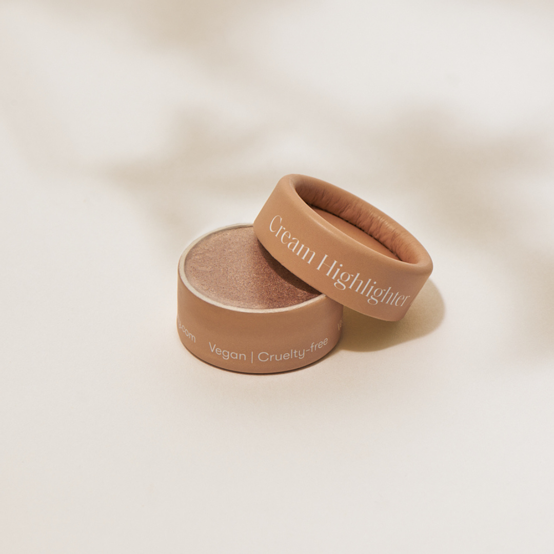 A small, round container of Minori’s Zero Waste Cream Highlighter Mini - Champagne with a brown matte lid sits at an angle; its packaging reads “Vegan, Cruelty-free,” reflecting clean beauty values on a softly lit neutral background.