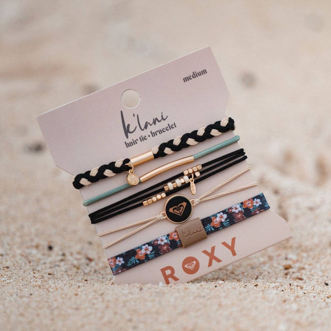 A set of six K’lani hair tie bracelets from the “Roxy Coastal Drift” collection, featuring black and neutral beachy designs with surf-inspired details, braided textures, and floral and pastel accents. Displayed on a branded card against sandy ground.