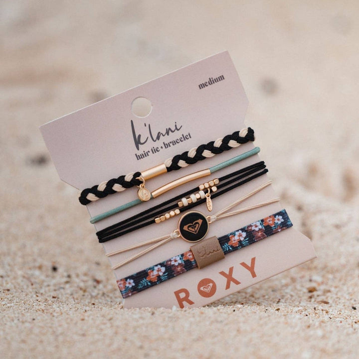 A set of six K’lani hair tie bracelets from the “Roxy Coastal Drift” collection, featuring black and neutral beachy designs with surf-inspired details, braided textures, and floral and pastel accents. Displayed on a branded card against sandy ground.