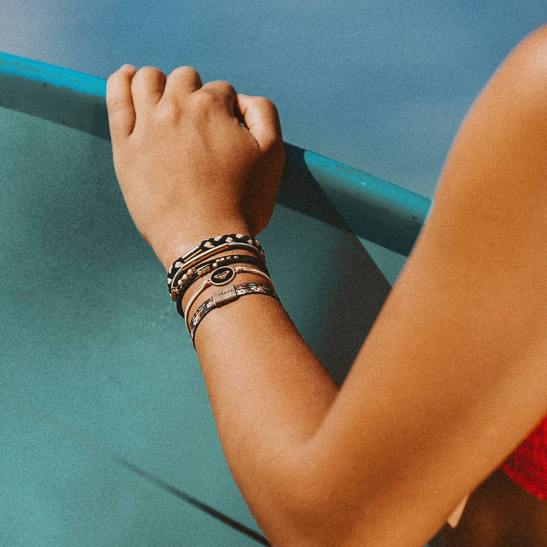 A person wearing multiple bracelets, including K’lani hair tie bracelets from the “Roxy Coastal Drift” Black &amp; Neutral Beachy Vibe set with braided textures, leans their arm on a turquoise surface as sunlight casts soft shadows against a backdrop of blue sky.