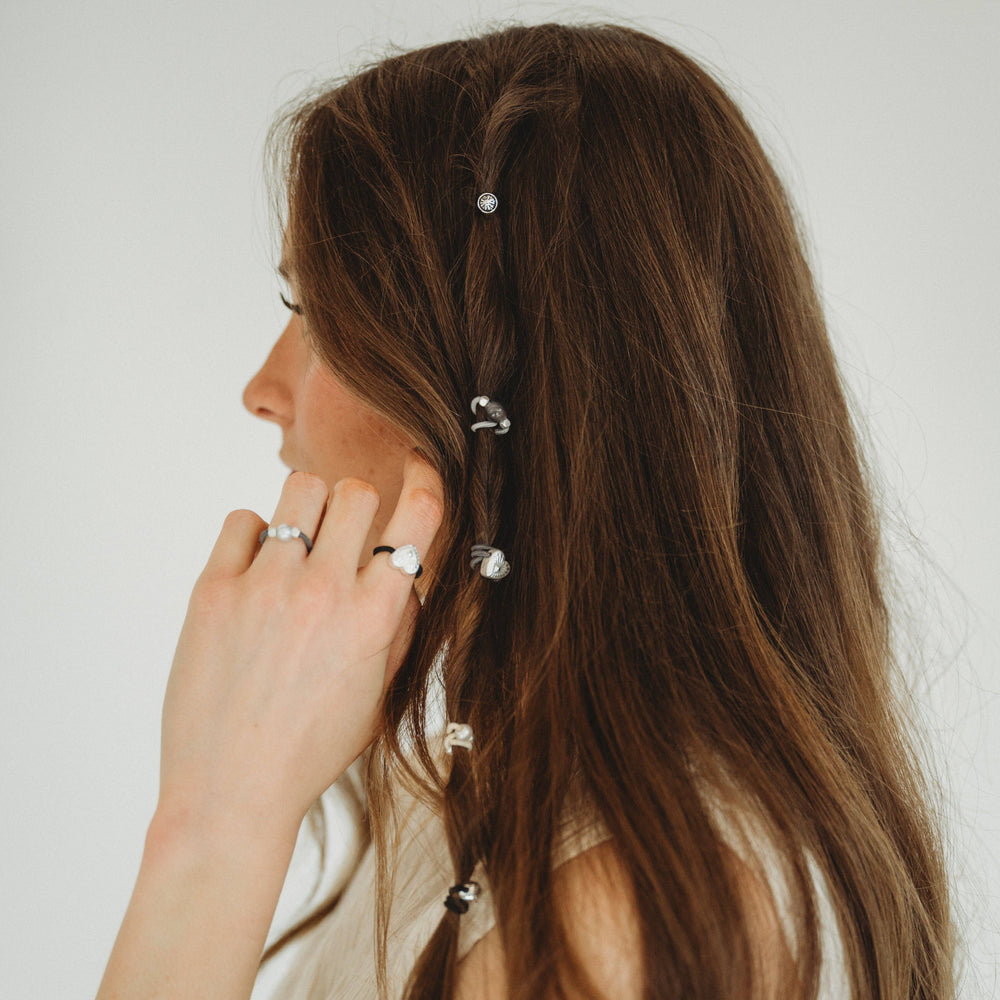 A person with long brown hair wears K’lani “Luna” Silver & Cool Tones Hair Tie Rings as silver accents, along with delicate pearl hair accessories braided into their hair for an elegant touch. The photo is taken from the side against a plain background.