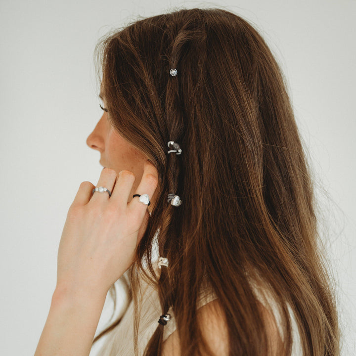A person with long brown hair wears K’lani “Luna” Silver &amp; Cool Tones Hair Tie Rings as silver accents, along with delicate pearl hair accessories braided into their hair for an elegant touch. The photo is taken from the side against a plain background.