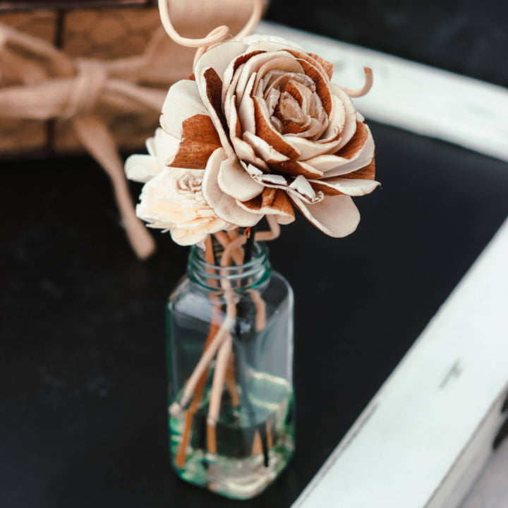 The WINTER &amp; HOLIDAY: Flower Reed Diffuser by Cedar Mountain Candle features light brown and cream-colored sola wood-inspired dried flowers on thin stems, elegantly arranged in a small glass bottle. The diffuser is displayed on a dark surface with a white frame and a burlap-wrapped box softly blurred in the background.