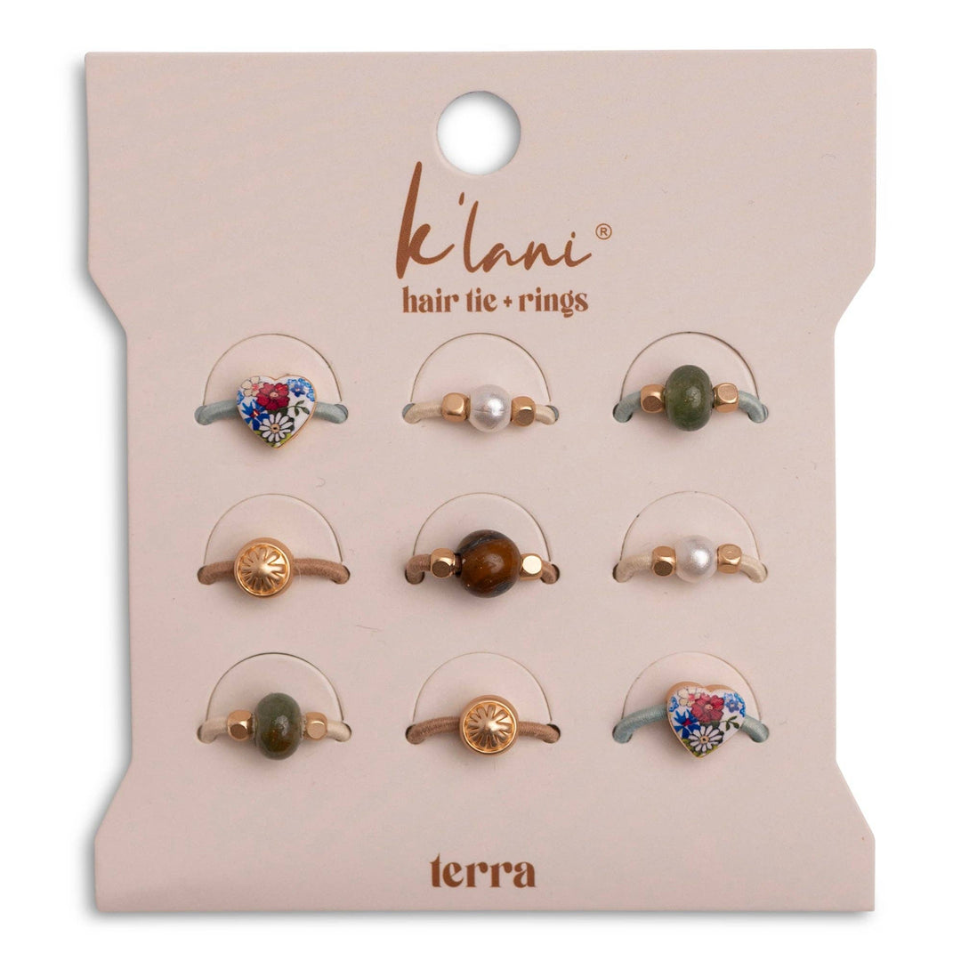 A set of nine versatile and decorative hair accessories—hair ties and hair tie rings—from K’lani hair tie bracelets, displayed on a beige card labeled “Terra” Earth Tones Hair Tie Rings. The collection features playful designs with hearts, pearls, beads, and gold accents.