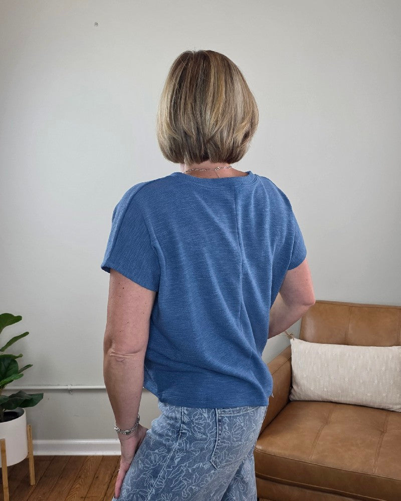 A person with short blonde hair, wearing the ZENANA Amanda Boxy Fit Cropped Tee with Pocket in blue and light patterned jeans, stands with their back to the camera in a living room featuring a tan sofa and a plant.
