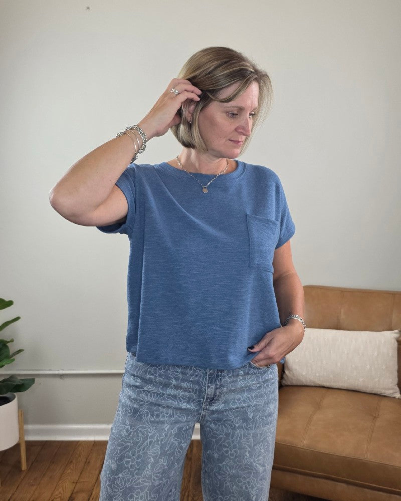 A woman wearing the ZENANA Amanda Boxy Fit Cropped Tee with Pocket in blue pairs it with patterned jeans as she stands indoors, looking down and touching her hair. A tan couch with pillows and a potted plant in the background highlight the tee’s relaxed, boxy silhouette.
