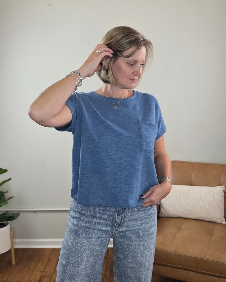 A woman wearing the ZENANA Amanda Boxy Fit Cropped Tee with Pocket in blue pairs it with patterned jeans as she stands indoors, looking down and touching her hair. A tan couch with pillows and a potted plant in the background highlight the tee’s relaxed, boxy silhouette.
