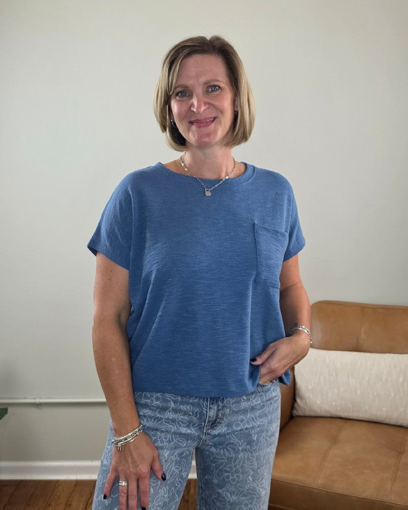 A woman with a blond bob haircut smiles while standing indoors. She wears the ZENANA Amanda Boxy Fit Cropped Tee with Pocket in blue, featuring a crew neckline, paired with patterned jeans for a relaxed silhouette. A tan couch and a pillow are visible in the background.