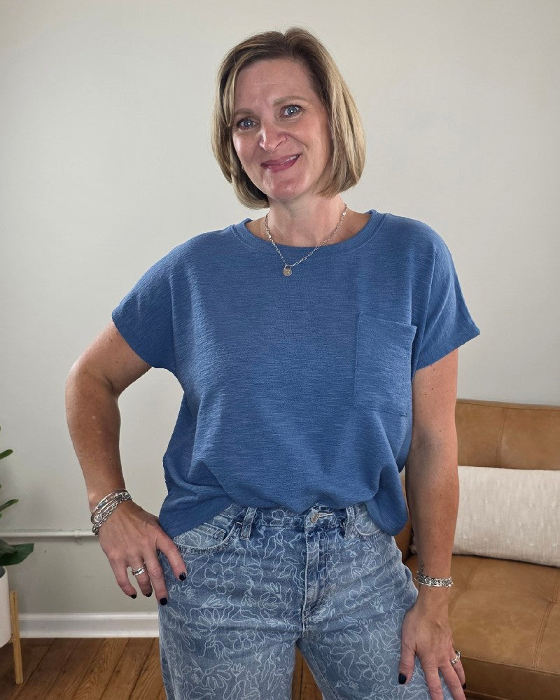 A smiling woman with short blonde hair stands indoors, wearing the ZENANA Amanda Boxy Fit Cropped Tee with Pocket in blue, paired with light blue patterned jeans. With one hand on her hip, she poses in front of a tan couch and a plant, highlighting the tee’s relaxed boxy silhouette and crew neckline.
