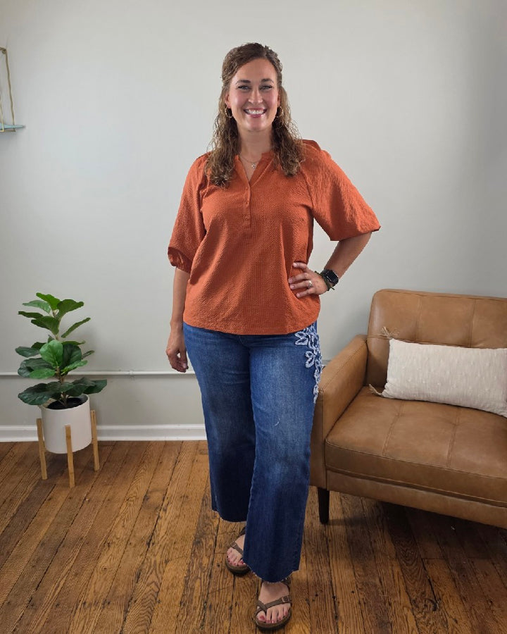 A smiling woman with curly hair wears the Heimish Amber Textured 3/4 Sleeve Blouse in a warm rust cinnamon hue, paired with wide-leg blue jeans featuring a floral design and sandals. She stands on a wooden floor near a tan couch and a potted plant against a light-colored wall.