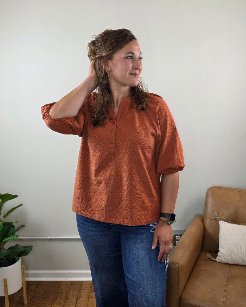 Wearing the Heimish Amber Textured 3/4 Sleeve Blouse and blue jeans, a woman stands in a living room, smiling as she touches her hair. She is positioned beside a brown leather chair and a green potted plant against a light-colored wall.