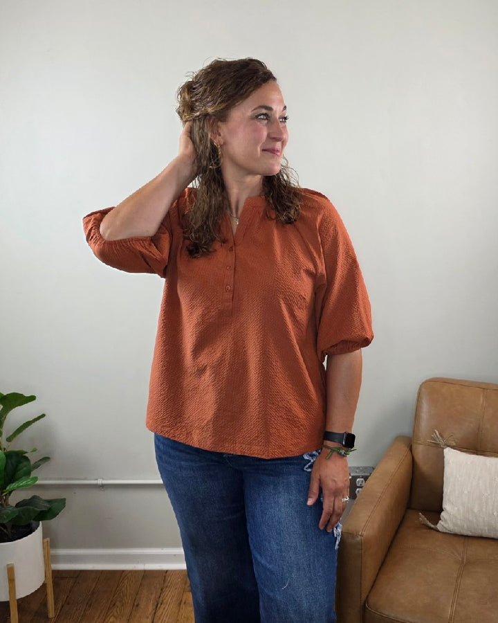 Wearing the Heimish Amber Textured 3/4 Sleeve Blouse and blue jeans, a woman stands in a living room, smiling as she touches her hair. She is positioned beside a brown leather chair and a green potted plant against a light-colored wall.