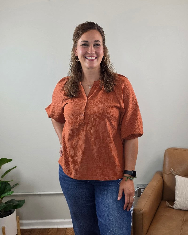A woman with curly brown hair, wearing the Heimish Amber Textured 3/4 Sleeve Blouse and blue jeans, smiles while standing in a living room with beige walls, a tan sofa, and a potted plant.