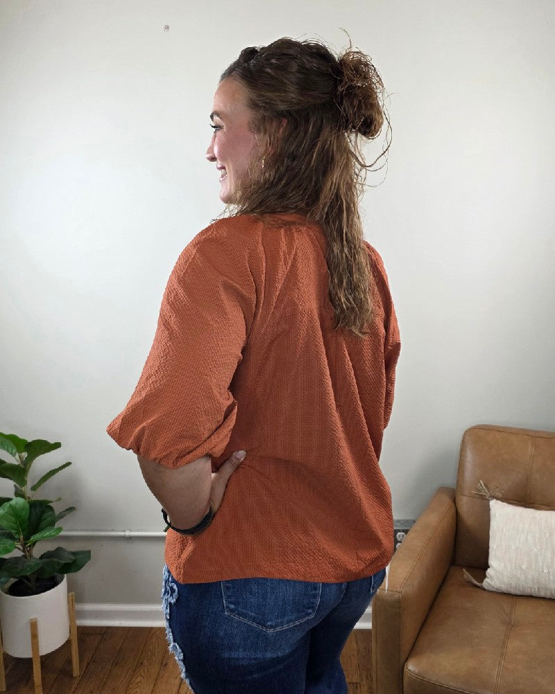 A woman with wavy brown hair stands sideways, smiling, wearing the Heimish Amber Textured 3/4 Sleeve Blouse and blue jeans. She is indoors near a tan couch and a potted plant against a light-colored wall.