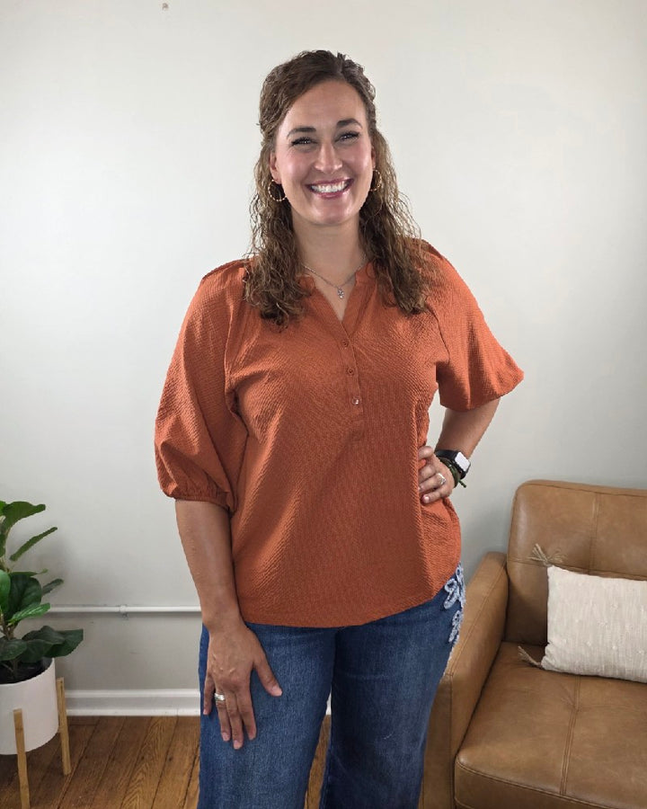 A woman with curly brown hair, dressed in the Heimish Amber Textured 3/4 Sleeve Blouse and blue jeans, stands smiling in a living room featuring a tan couch and a potted plant against a light-colored wall.