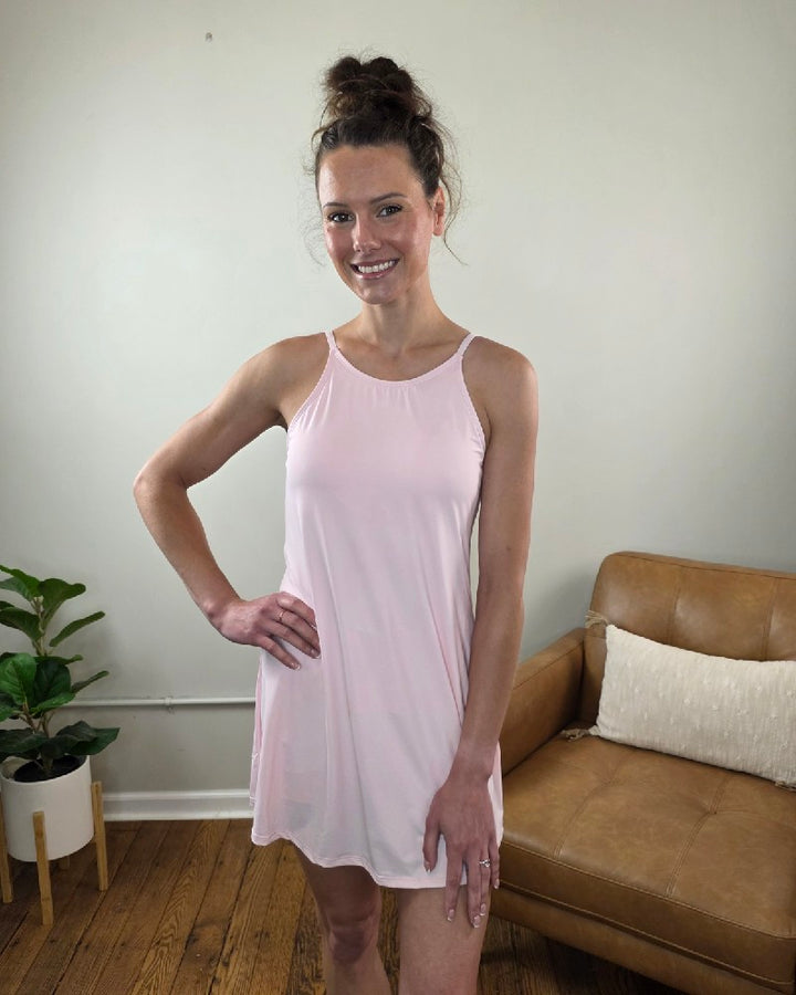A woman smiles indoors wearing the Shewin Amelia Tank Dress with Athletic Shorts in light pink, her hair in a bun. She stands on a wooden floor near a tan couch and a green potted plant.