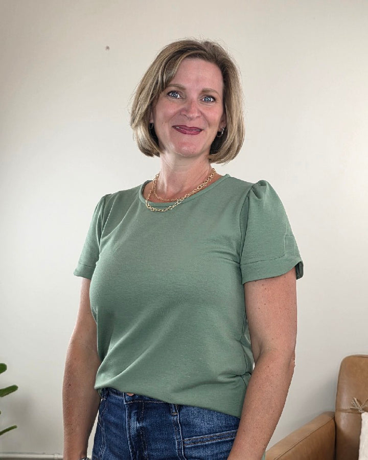 A woman with short blonde hair, wearing the Les Amis Arlet Tulip Sleeve Blouse in green and jeans, stands indoors smiling at the camera. A tan couch and a green plant are visible in the background.