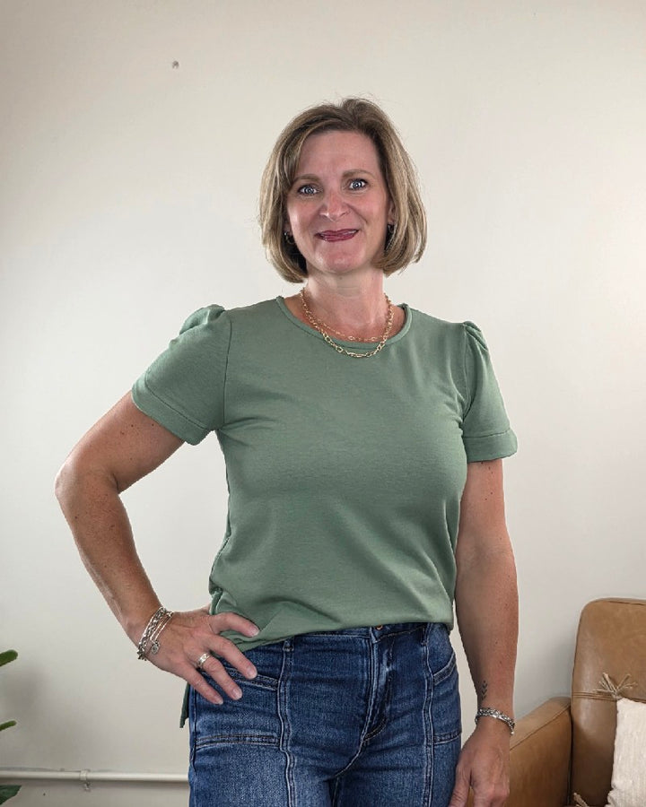 A woman with short blonde hair smiles at the camera, wearing the Les Amis Arlet Tulip Sleeve Blouse in green made from medium-weight fabric. She has paired it with blue jeans and jewelry, and is standing indoors in front of a beige wall and a brown chair.