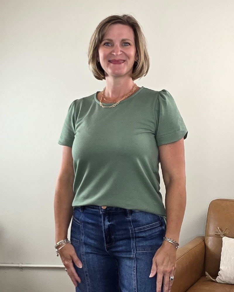 A woman with short blonde hair is wearing the Les Amis Arlet Tulip Sleeve Blouse in green, made from a medium weight fabric, paired with jeans as she stands smiling in a room with a beige couch and a white wall behind her.