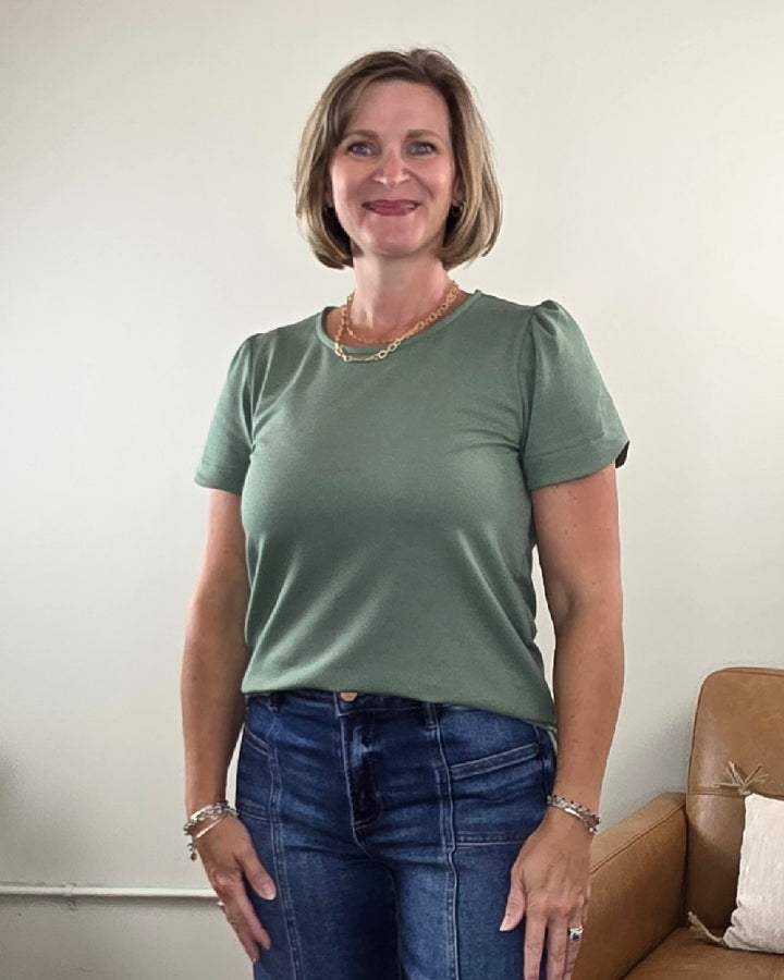 A woman with short blonde hair is wearing the Les Amis Arlet Tulip Sleeve Blouse in green, made from a medium weight fabric, paired with jeans as she stands smiling in a room with a beige couch and a white wall behind her.