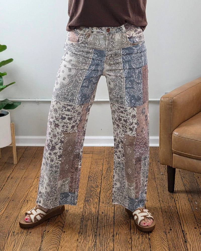 A person wearing the Aven High Rise Ankle Wide Leg Jeans from Risen, paired with a brown shirt and white sandals, stands on a wooden floor. Nearby are a potted plant and a tan armchair set against a white wall.