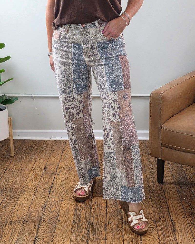 A person stands indoors wearing the Risen Aven High Rise Ankle Wide Leg Jeans, styled with a brown top and white sandals. Part of a brown chair, a plant, and wood flooring are visible in the background.