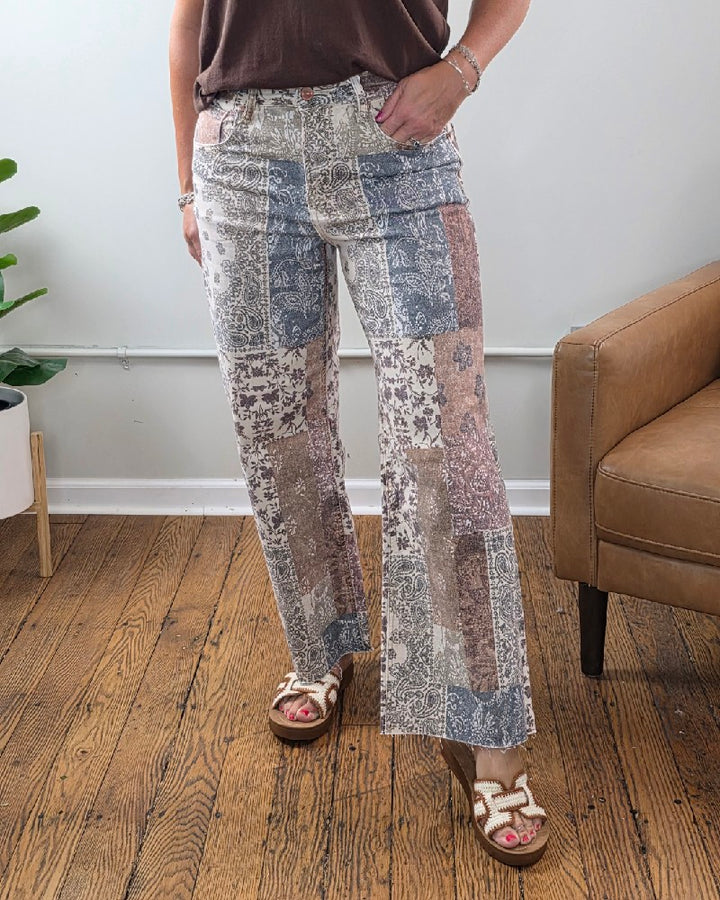 A person stands indoors wearing the Risen Aven High Rise Ankle Wide Leg Jeans, styled with a brown top and white sandals. Part of a brown chair, a plant, and wood flooring are visible in the background.