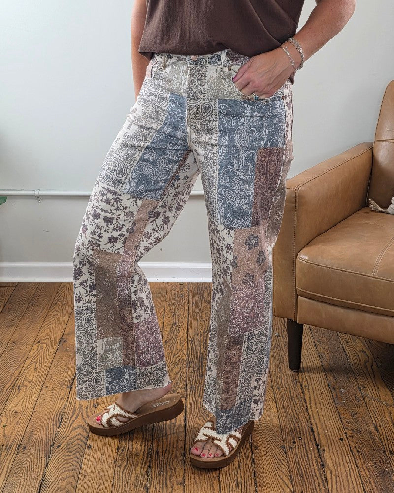 A person stands on a wooden floor next to a tan armchair, wearing Risen’s Aven High Rise Ankle Wide Leg Jeans and sandals. Only the lower half of their body is visible, with one hand resting in a pocket.