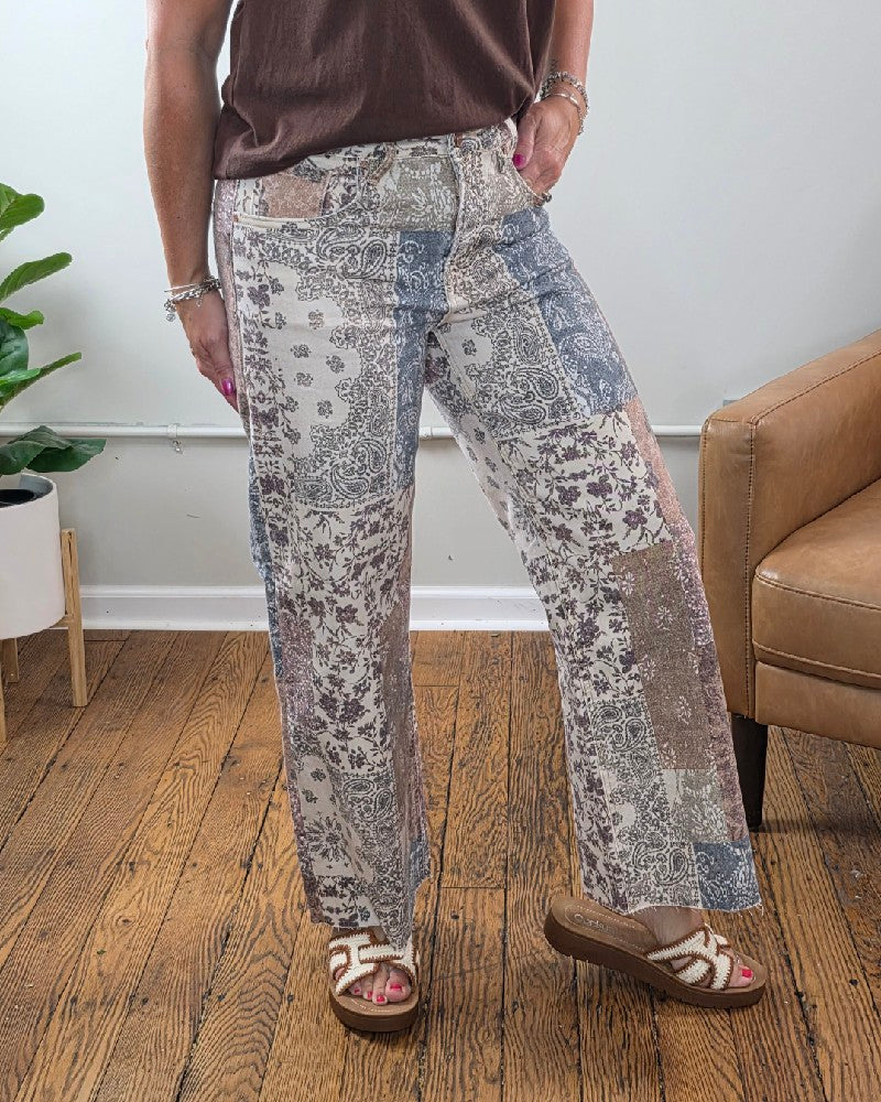 A person wearing the Aven High Rise Ankle Wide Leg Jeans from Risen, a brown top, and sandals stands on a wooden floor near a tan chair and a green potted plant.