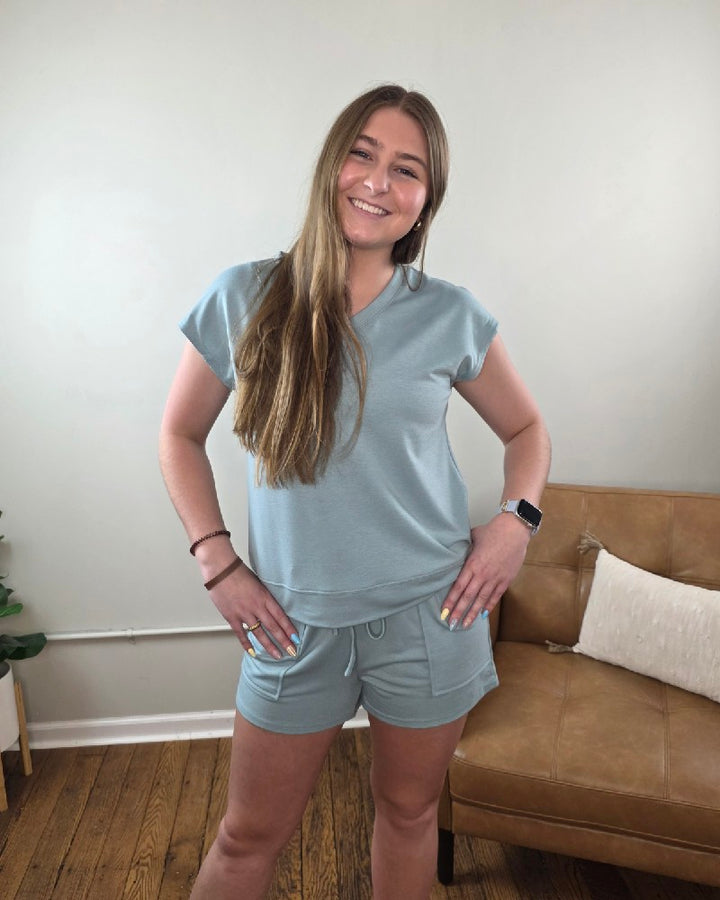 A young woman with long hair smiles indoors, wearing the Lovesoft Avery V-Neck Top and Shorts Lounge Set in light blue. A tan couch with a pillow and wooden floor are seen in the background.
