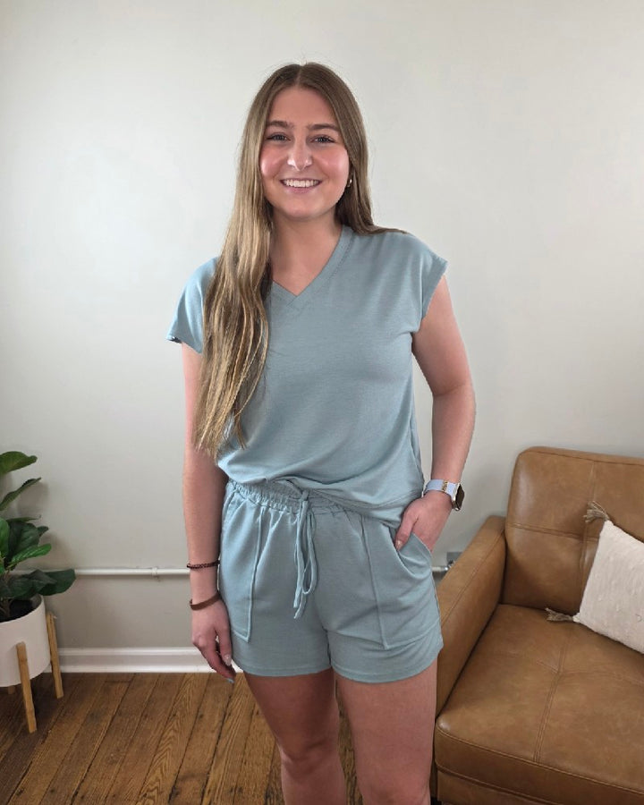 A woman with long brown hair smiles indoors, wearing the Lovesoft Avery V-Neck Top and Shorts Lounge Set in light blue. A tan couch and a plant are visible in the background.