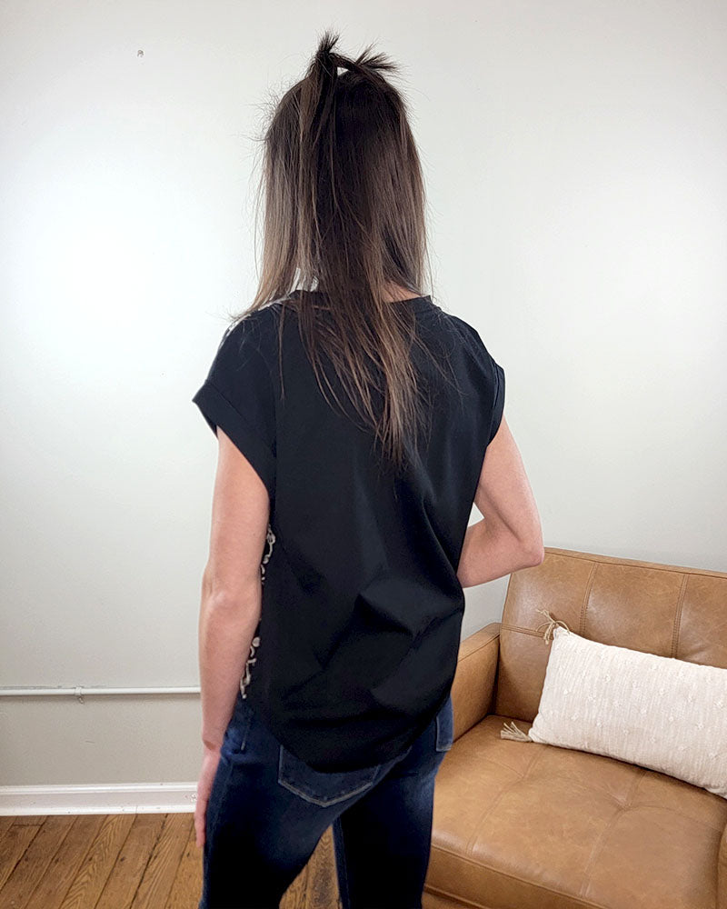 A person with long brown hair wears the Staccato Rita Embroidered Blouse, a black short-sleeve top with delicate floral embroidery, and dark jeans, standing with their back to the camera near a tan leather couch and white pillow.