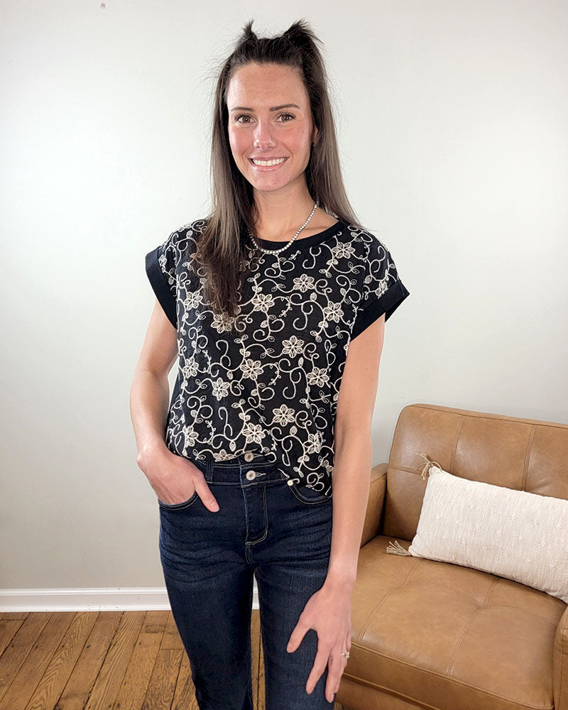 A woman with long brown hair smiles at the camera indoors, wearing the Staccato Rita Embroidered Blouse—a black short-sleeve top with delicate floral embroidery—and dark jeans. She stands beside a tan chair with a white pillow, one hand in her pocket.