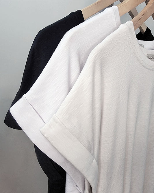 Three Thea Rolled Sleeve Tees by ZENANA—two white and one black—crafted from a soft cotton blend, hang on wooden hangers against a light gray background. These tees are ideal closet staples for any wardrobe.