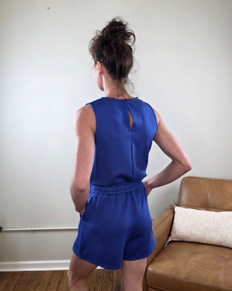 Person wearing the Blaise Sleeveless Romper in blue standing in a room with a brown couch and white wall.