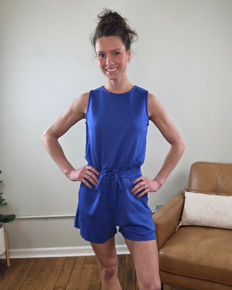 Woman wearing the Blaise Sleeveless  blue romper standing in a room with a couch and plant.