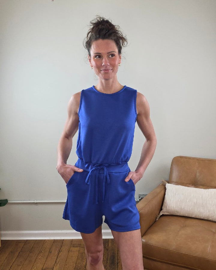 Woman wearing the Blaise Sleeveless Romper in blue standing in a room with a brown couch.