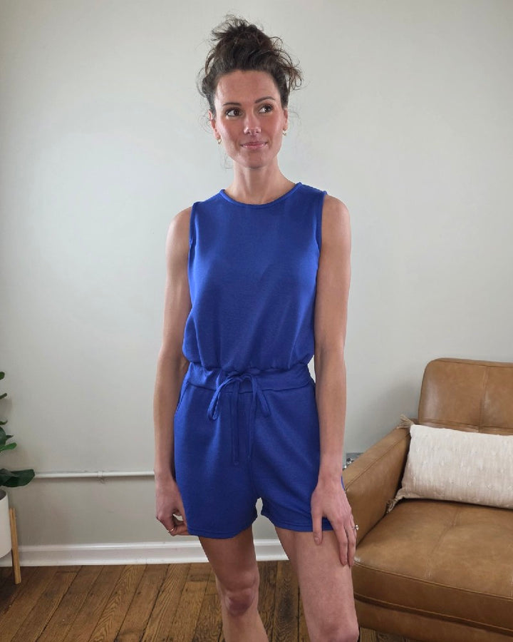 Woman wearing the Blaise Sleeveless Romper in blue standing in a room with a brown couch and white wall.