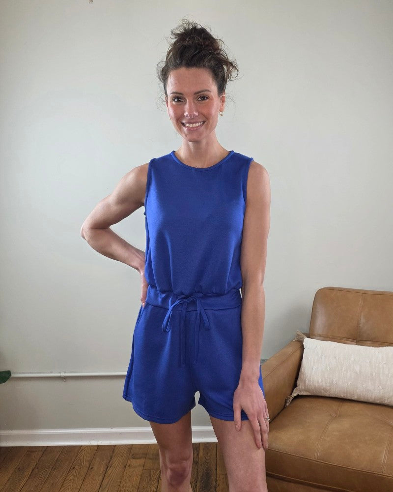 Woman wearing the Blaise Sleeveless Romper in blue standing in a room with a beige wall and brown couch.