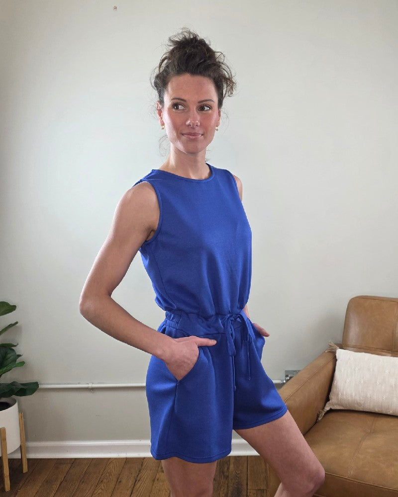 Woman wearing the Blaise Sleeveless Romper in blue standing in a room with a white wall and brown chair.
