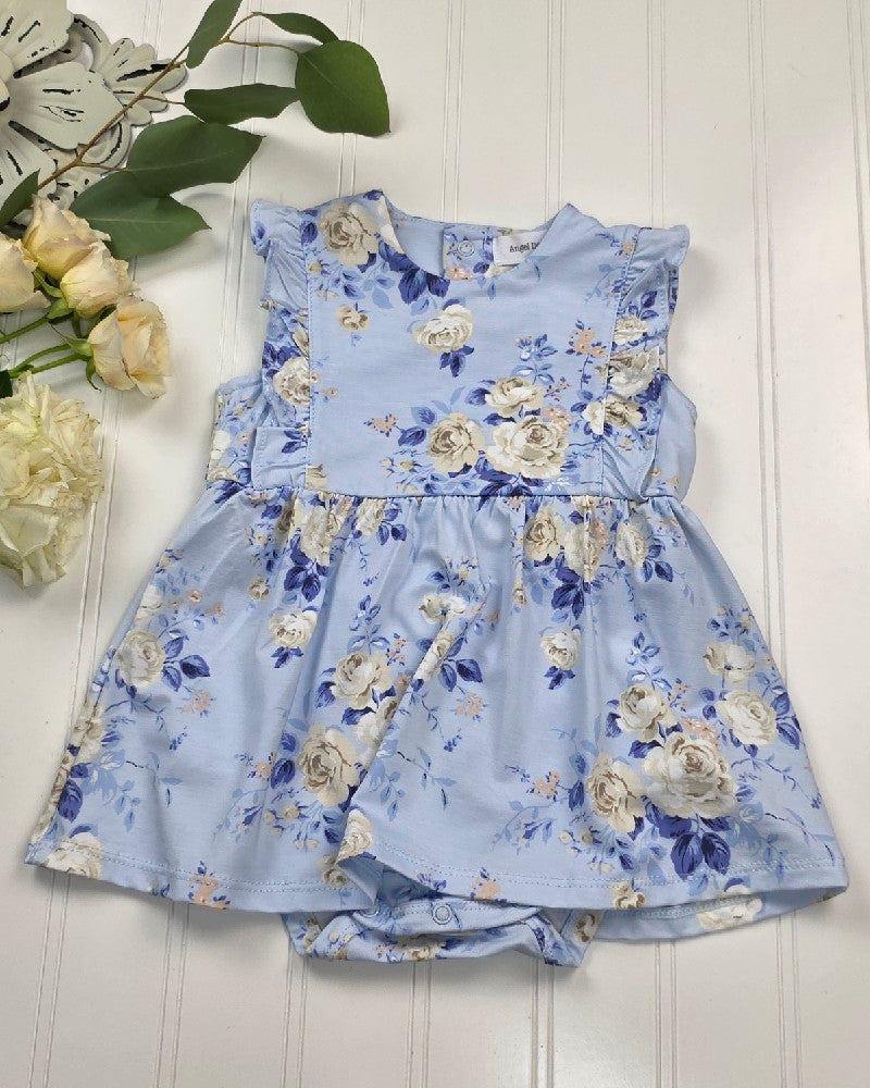 The Ashley Rose Floral - Ruffle Bodysuit Dress by Angel Dear, featuring white and blue floral prints, ruffle sleeves, and a gathered skirt, is displayed on a white wooden surface with cream roses and green leaves nearby.