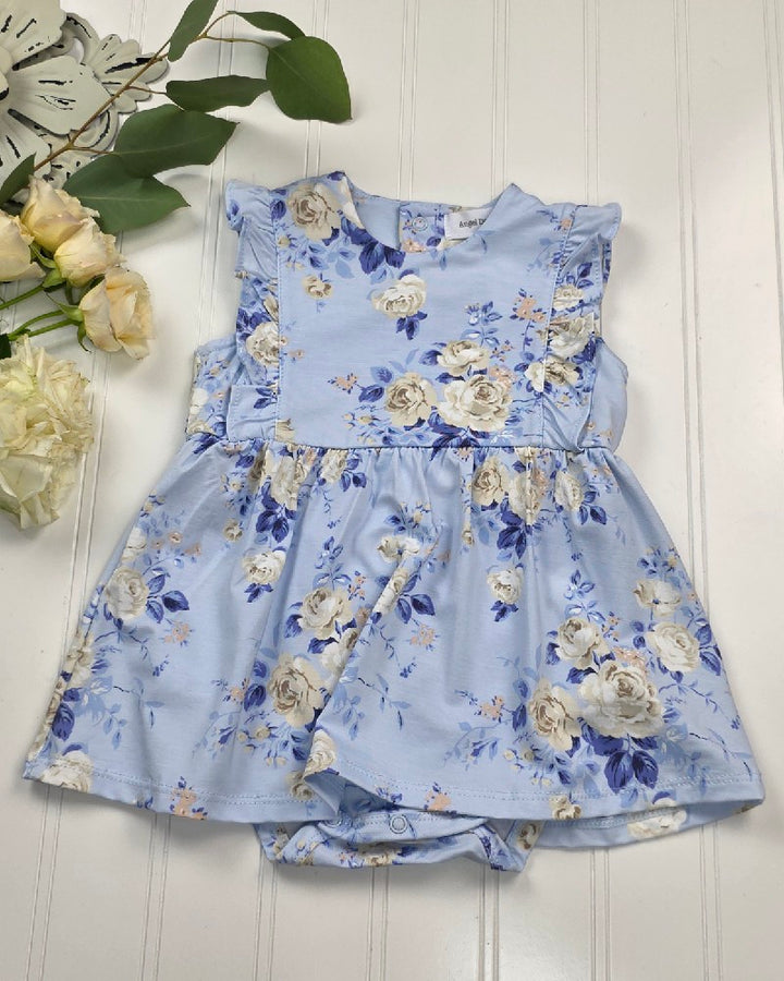 The Ashley Rose Floral - Ruffle Bodysuit Dress by Angel Dear, featuring white and blue floral prints, ruffle sleeves, and a gathered skirt, is displayed on a white wooden surface with cream roses and green leaves nearby.