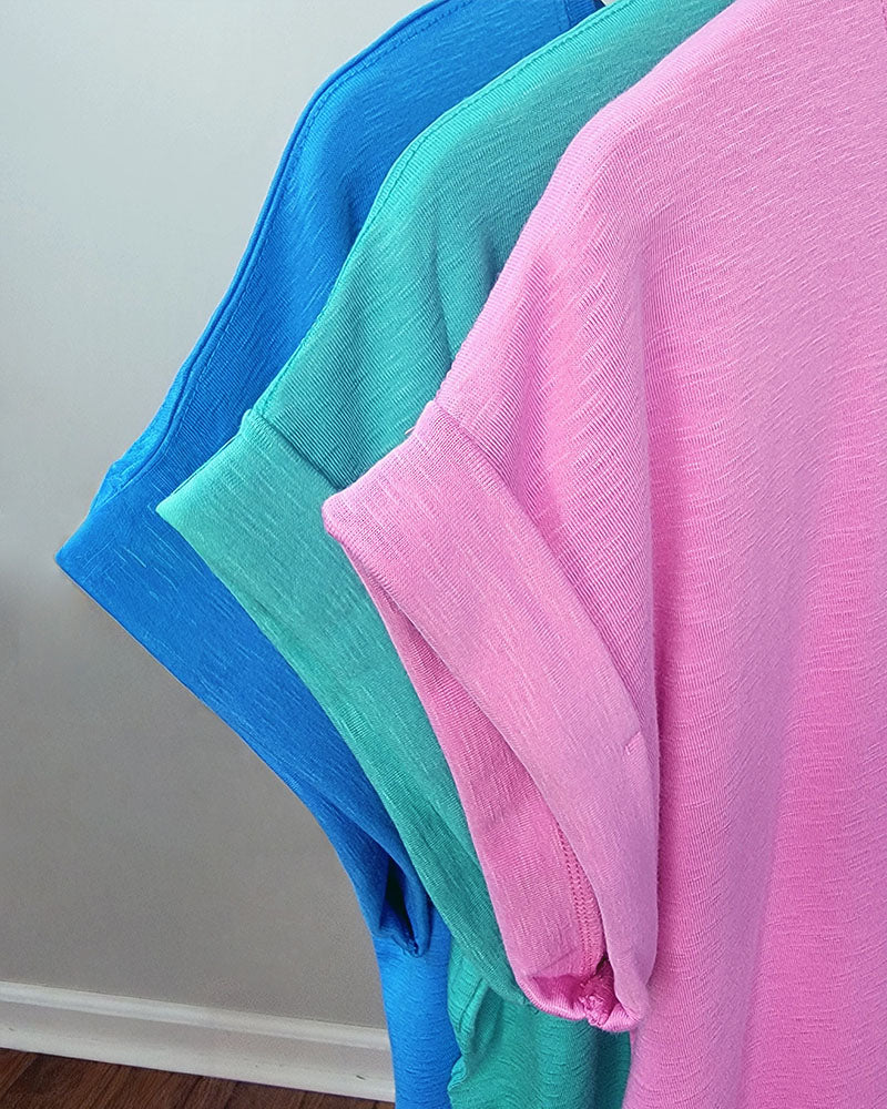 Three ZENANA Thea Rolled Sleeve Tees in pink, green, and blue hang side by side on hangers against a light wall and wood floor. These tees feature soft, textured cotton-blend fabric for added comfort.