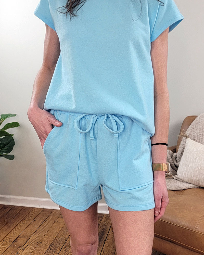 A woman models the Mono B Reagan Set, a blue two-piece outfit with elastic drawstring shorts, indoors. She stands with one hand in her pocket; a wooden floor, plant, and brown sofa with cushions are in the background.
