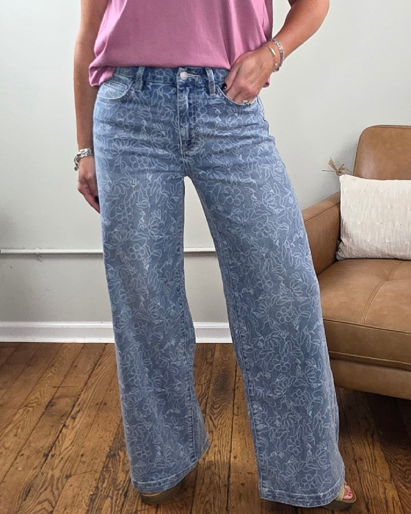 A person wearing the Briella Floral Print, High Rise, Wide Leg Jeans from Vervet paired with a pink top stands on a wooden floor near a brown armchair and a white wall. The high rise design of these floral print jeans by Vervet adds a stylish touch to the denim look.