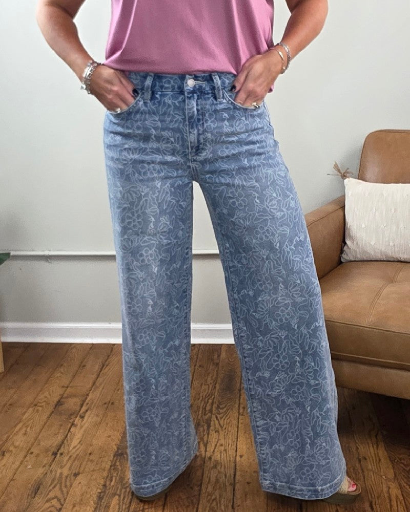 Wearing Vervet’s Briella Floral Print High Rise Wide Leg Jeans, paired with a pink short-sleeve top and bracelets, the person stands indoors on a wooden floor next to a tan chair with a white pillow.
