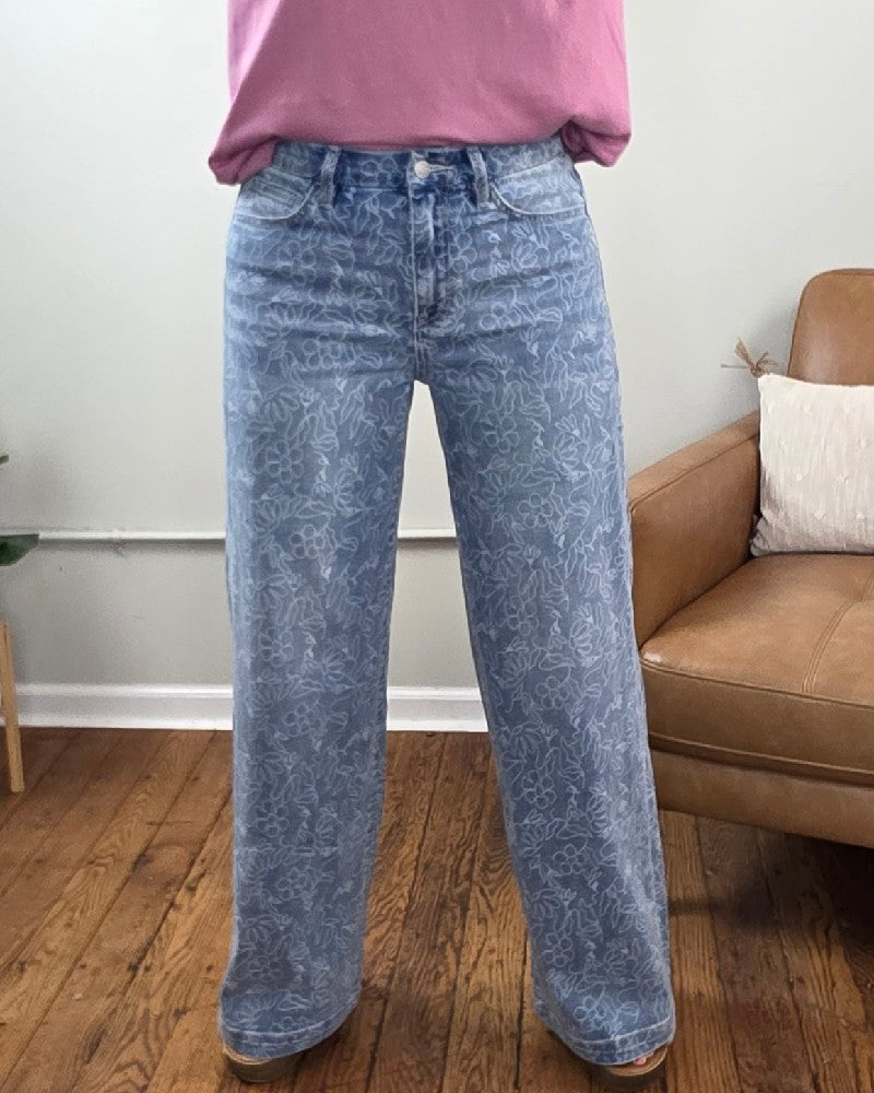 A person wearing the Briella Floral Print, High Rise, Wide Leg Jeans from Vervet paired with a pink shirt is standing indoors on a wooden floor next to a brown chair with a white pillow.