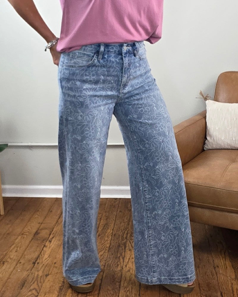 A person wearing Vervet’s Briella Floral Print High Rise Wide Leg Jeans paired with a pink t-shirt, standing on a wooden floor next to a tan chair.