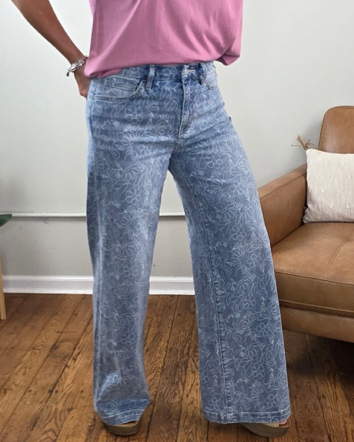 A person wearing Vervet’s Briella Floral Print High Rise Wide Leg Jeans paired with a pink t-shirt, standing on a wooden floor next to a tan chair.
